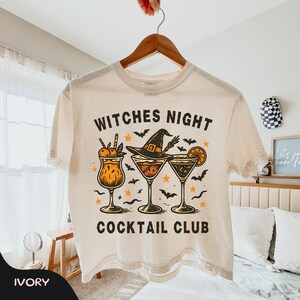 May include: An ivory short-sleeve t-shirt featuring a graphic print with the text "WITCHES NIGHT COCKTAIL CLUB". The design includes three cocktail glasses, a witch hat, small stars, and bats in shades of orange, black, and yellow.