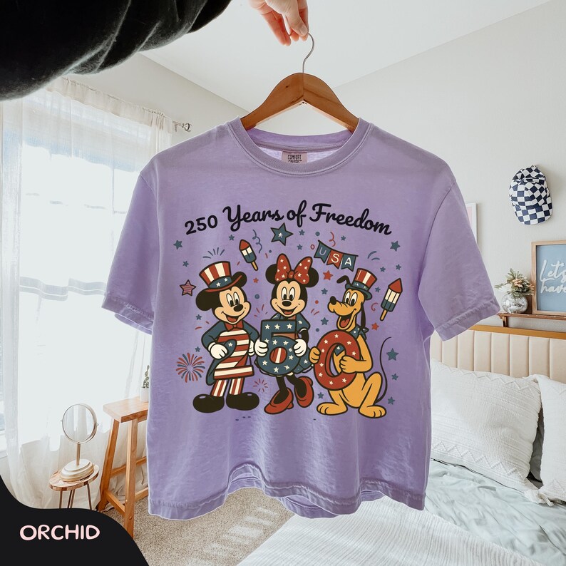 Może przedstawiać: Liliowa koszulka z Myszka Miki, Myszka Minnie i Pluto w patriotycznych strojach. Koszulka ma napis "250 Years of Freedom USA" i wisi na drewnianym wieszaku.