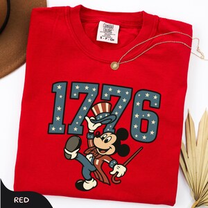 May include: A red t-shirt featuring the number "1776" in blue with white stars, and a cartoon character in patriotic attire. The shirt has a crew neck and a small tag on the upper back. A gold necklace is draped across the shirt.