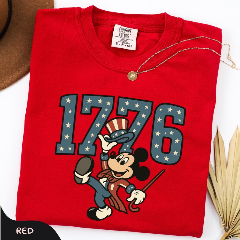 May include: A red t-shirt featuring the number "1776" in blue with white stars, and a cartoon character in patriotic attire. The shirt has a crew neck and a small tag on the upper back. A gold necklace is draped across the shirt.