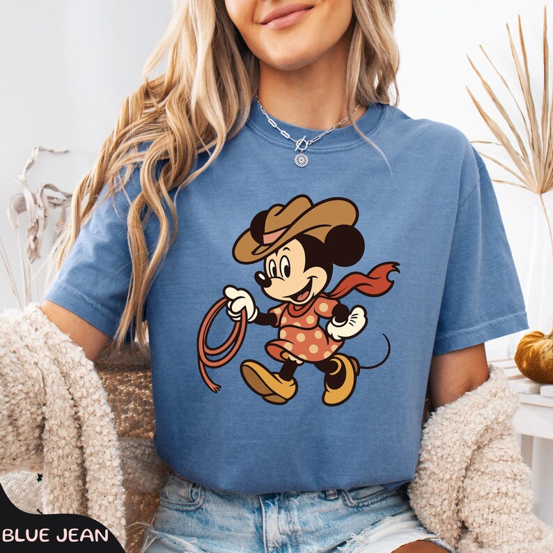 May include: A blue jean t-shirt featuring a cartoon Minnie Mouse dressed as a cowgirl, wearing a brown hat, a red scarf, and a polka-dot dress. She is holding a lasso. The shirt has short sleeves.