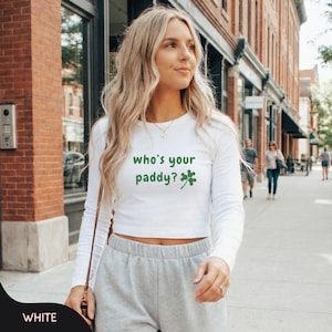 May include: White long-sleeved crop top featuring the green text "who's your paddy?" and a shamrock. The top is worn with grey sweatpants. The image is taken outside.