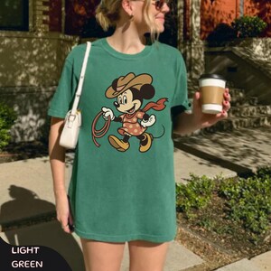 May include: Light green t-shirt featuring a cartoon Minnie Mouse dressed as a cowgirl, holding a lasso. The design includes a brown cowboy hat, a red scarf, and a polka-dot dress. The words "LIGHT GREEN" are at the bottom of the image.