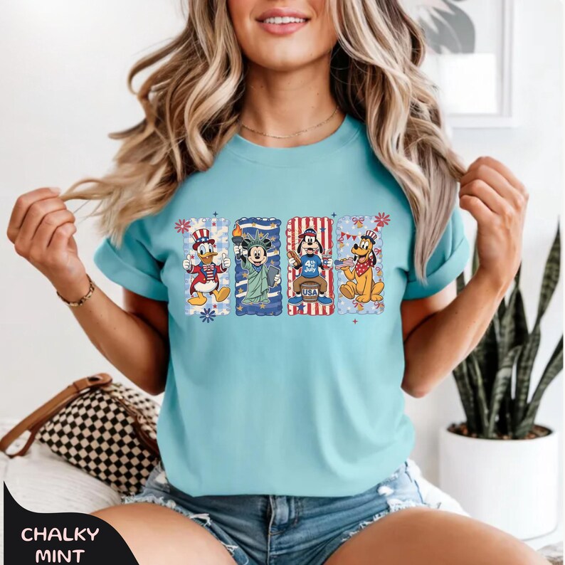 May include: A light blue t-shirt featuring four cartoon characters in patriotic designs. Donald Duck, Mickey Mouse, Goofy, and Pluto are depicted in red, white, and blue, with the text "USA" on Goofy's design. The shirt has the text "CHALKY MINT" at the bottom.