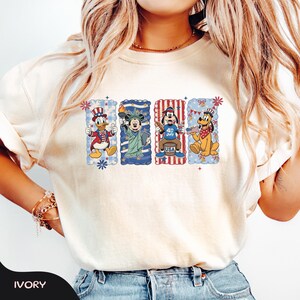 May include: Ivory-colored t-shirt featuring four cartoon characters in patriotic designs. Donald Duck, Mickey Mouse, Goofy, and Pluto are depicted with American flag motifs and 4th of July themes. The shirt has a crew neck and short sleeves.