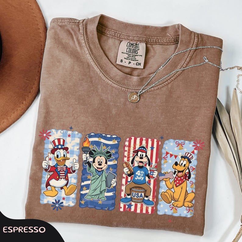 May include: Espresso-colored t-shirt featuring four cartoon characters in patriotic designs. Donald Duck, Mickey Mouse, Goofy, and Pluto are depicted in red, white, and blue, celebrating the 4th of July. A silver necklace is draped across the shirt.