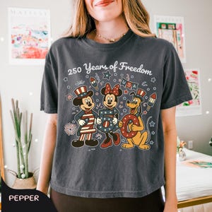 Może przedstawiać: Ciemnoszara koszulka z Myszką Miki, Myszka Minnie i Pluto w patriotycznych strojach. Napis "250 Years of Freedom" i elementy USA w kolorach czerwonym, białym i niebieskim. Styl vintage.