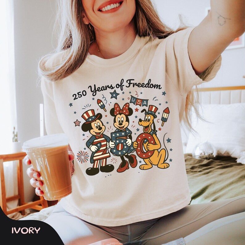 Może przedstawiać: Kremowa koszulka z napisem "250 Years of Freedom" i grafiką przedstawiającą Myszki Miki, Minnie i Pluto w patriotycznych strojach. Projekt zawiera gwiazdy, fajerwerki i słowo "USA".