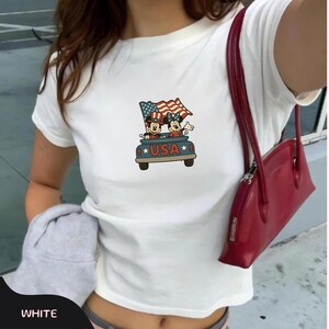 May include: White t-shirt featuring a graphic of Mickey and Minnie Mouse in a car with a waving American flag. The car has the word "USA" on it. The shirt has short sleeves and a crew neck.