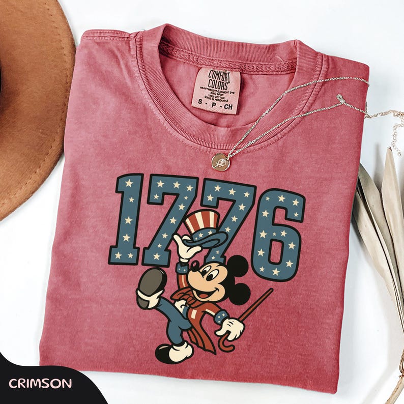 May include: Crimson-colored t-shirt featuring the number "1776" in blue with white stars, and a cartoon character dressed in patriotic attire. The shirt has a Comfort Colors tag and a silver necklace.