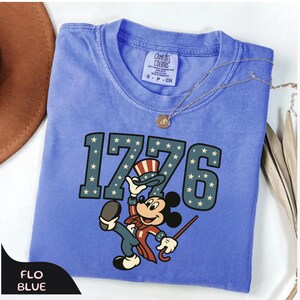 May include: A blue Comfort Colors t-shirt with the number "1776" in a star-spangled font. A cartoon mouse in patriotic attire is featured below the numbers. A gold necklace is draped across the shirt. The shirt is labeled "Flo Blue".
