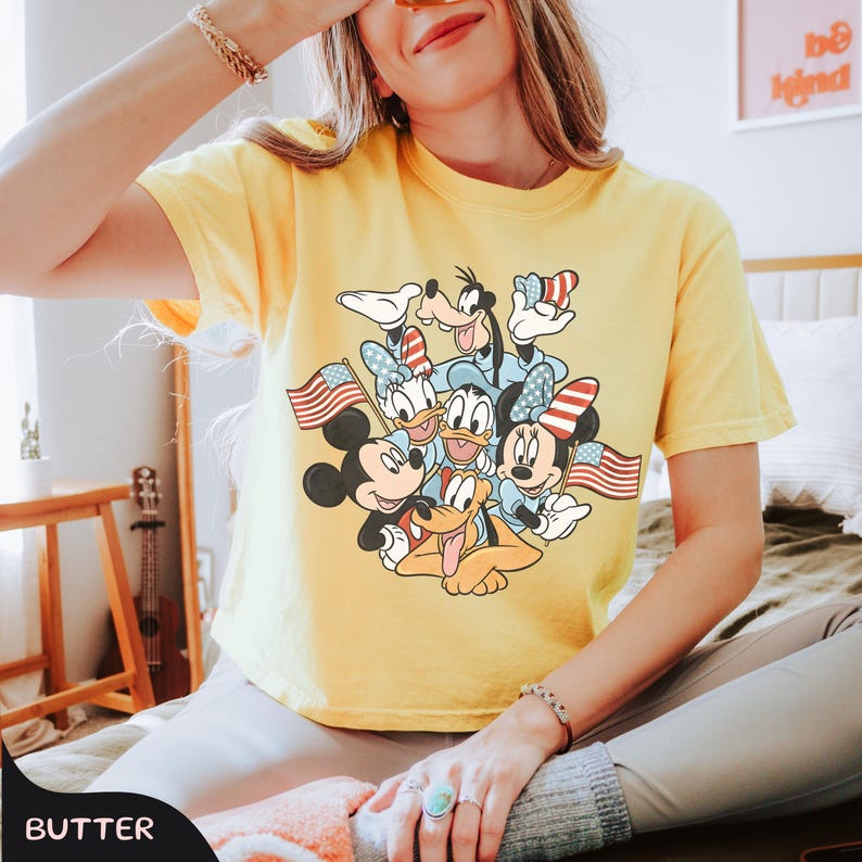 May include: Yellow t-shirt featuring a graphic of Disney characters Mickey Mouse, Minnie Mouse, Donald Duck, Daisy Duck, Goofy, and Pluto, holding American flags. The shirt has a crew neck and short sleeves.