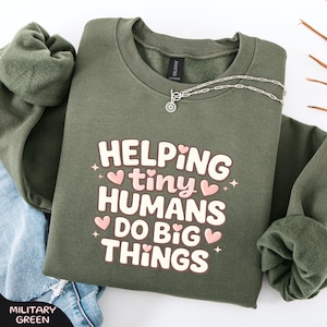 May include: A military green sweatshirt featuring the text "HELPING tiny HUMANS DO BIG THINGS" in white and pink. The sweatshirt has a crew neck and long sleeves. A silver necklace with a sun pendant is on the sweatshirt.