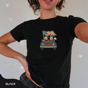 May include: Black t-shirt featuring a graphic of Mickey and Minnie Mouse in a car with American flags. The car has the word "USA" on it. The shirt is a comfortable, casual style.