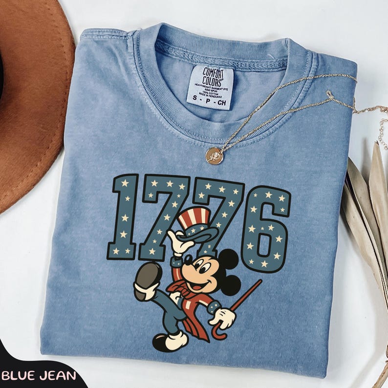 May include: A blue jean colored t-shirt featuring the number "1776" in a star-studded font, with a cartoon character dressed in patriotic attire. The shirt has a crew neck and short sleeves. A gold necklace with a pendant is draped across the shirt.