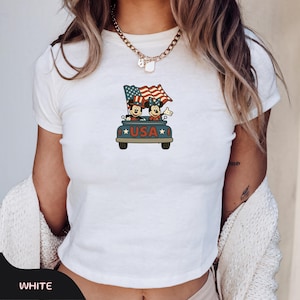 May include: White t-shirt featuring a cartoon graphic of Mickey and Minnie Mouse in a car with a waving American flag. The car has the word "USA" on it. The shirt is a crew neck style.
