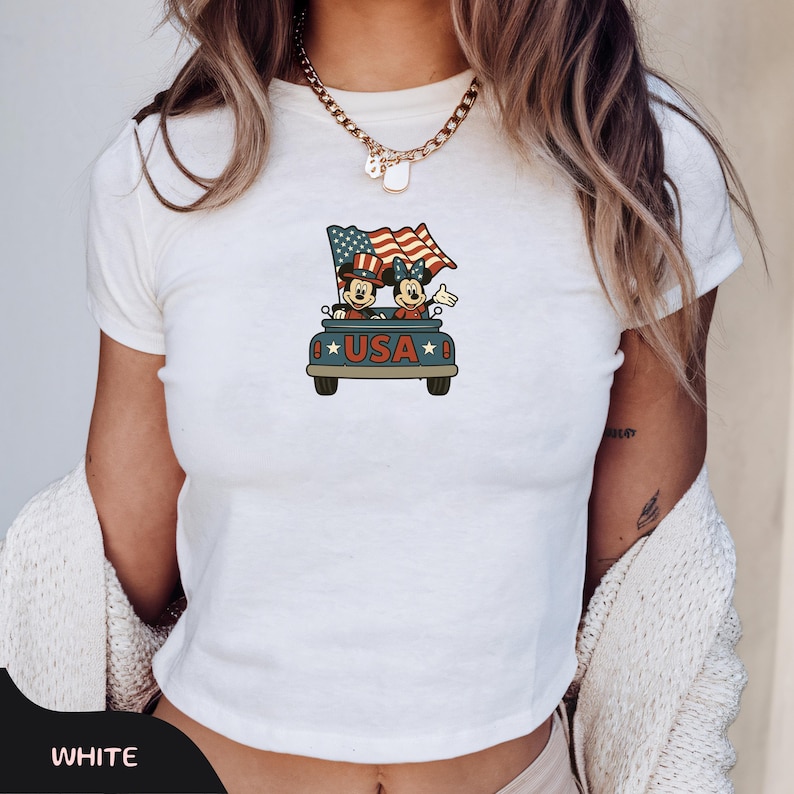 May include: White t-shirt featuring a cartoon graphic of Mickey and Minnie Mouse in a car with a waving American flag. The car has the word "USA" on it. The shirt is a crew neck style.