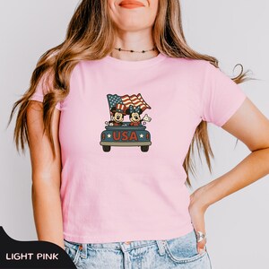 May include: Light pink t-shirt featuring a cartoon graphic of Mickey and Minnie Mouse in a car with a waving American flag. The car has the word "USA" on it. The shirt is a crew neck and short-sleeved.