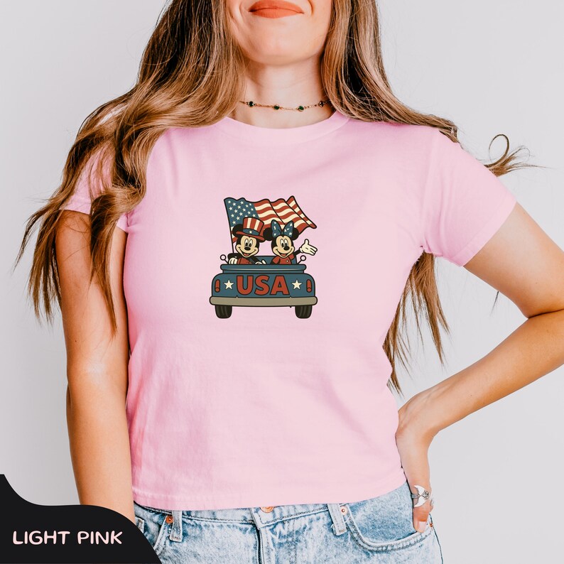 May include: Light pink t-shirt featuring a cartoon graphic of Mickey and Minnie Mouse in a car with a waving American flag. The car has the word "USA" on it. The shirt is a crew neck and short-sleeved.