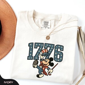 May include: Ivory-colored t-shirt featuring the number "1776" in blue with white stars, and a cartoon character in patriotic attire. The shirt has a crew neck and short sleeves. A gold necklace and a brown hat are also visible.