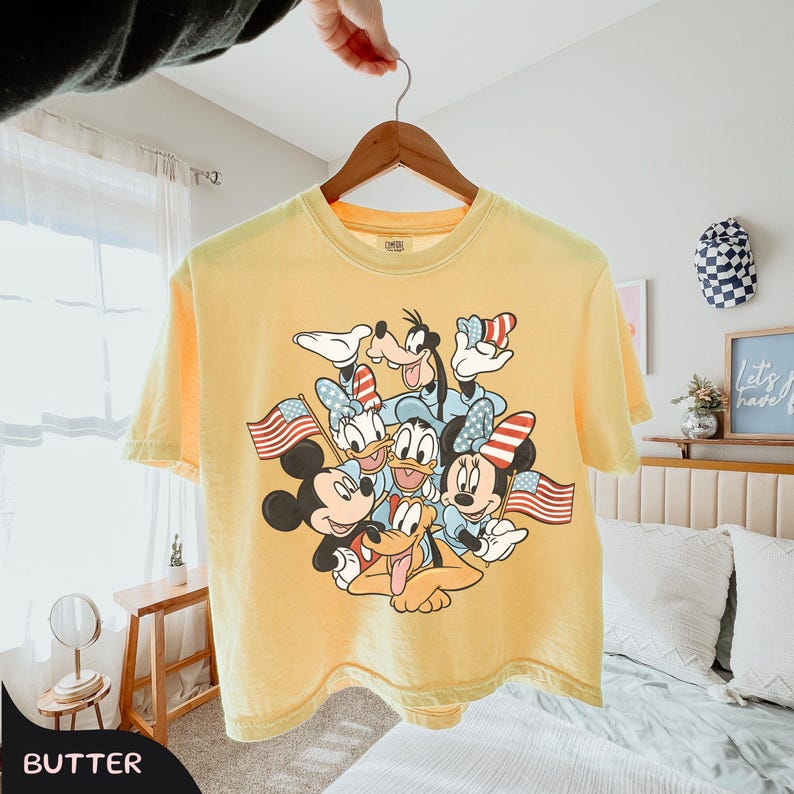 May include: Yellow short-sleeved t-shirt featuring a graphic of Mickey Mouse, Minnie Mouse, Donald Duck, Daisy Duck, Goofy, and Pluto, each holding an American flag. The shirt is hanging from a wooden hanger.