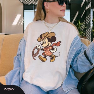May include: Ivory-colored t-shirt featuring a cartoon Minnie Mouse dressed as a cowgirl, wearing a brown hat, red scarf, and a pink polka-dot dress. She is holding a lasso. The word "IVORY" is printed at the bottom.