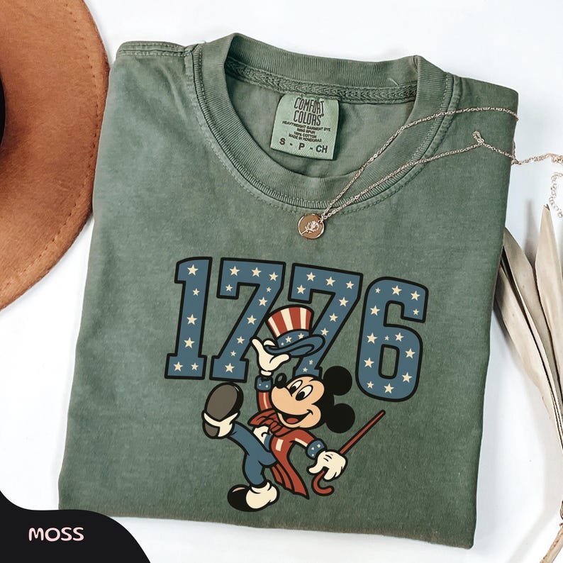 May include: A moss green t-shirt featuring the number "1776" in blue with white stars. A cartoon character in patriotic attire is below the numbers. The shirt has a crew neck and a small tag at the neckline. A gold necklace is draped across the shirt.