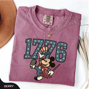 May include: Berry-colored t-shirt with the number "1776" in blue with white stars. A cartoon mouse in patriotic attire is featured below the numbers. The shirt has a Comfort Colors tag and a gold necklace. A brown hat is visible in the upper left corner.