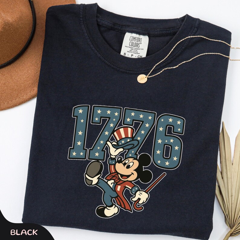 May include: A navy blue Comfort Colors t-shirt featuring a graphic of Mickey Mouse in patriotic attire, with the number "1776" in a star-spangled design. A gold necklace and a brown hat are also visible.