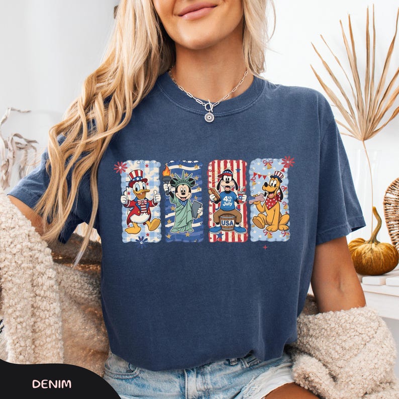 May include: Denim blue t-shirt featuring four cartoon characters in patriotic designs. Donald Duck in a red, white, and blue outfit, Mickey Mouse holding the Statue of Liberty torch, Goofy with a "4th of July" design, and Pluto with a red bandana.