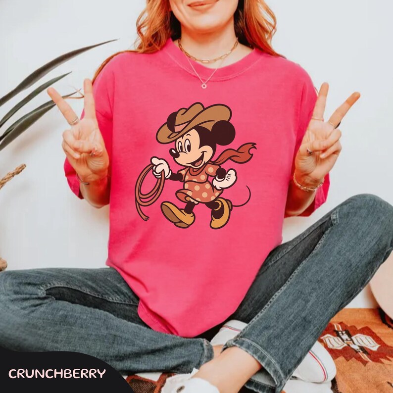 May include: A hot pink t-shirt featuring a cartoon Minnie Mouse dressed as a cowgirl, holding a lasso. She wears a brown cowboy hat, a red scarf, and a polka-dot dress. The word "CRUNCHBERRY" is at the bottom.