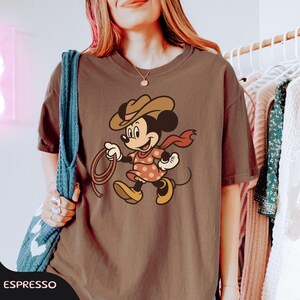 May include: Espresso-colored t-shirt featuring a cartoon Minnie Mouse dressed as a cowgirl. Minnie wears a brown hat, red scarf, and a pink dress with orange polka dots. She holds a lasso and is walking forward. The word "ESPRESSO" is at the bottom.