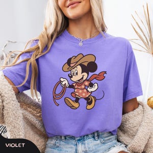 May include: Violet-colored t-shirt featuring a cartoon Minnie Mouse dressed as a cowgirl, wearing a brown hat, red scarf, and a pink dress with white polka dots. Minnie is holding a lasso. The word "VIOLET" is at the bottom.