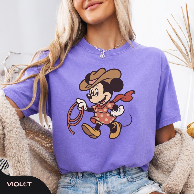 May include: Violet-colored t-shirt featuring a cartoon Minnie Mouse dressed as a cowgirl, wearing a brown hat, red scarf, and a pink dress with white polka dots. Minnie is holding a lasso. The word "VIOLET" is at the bottom.