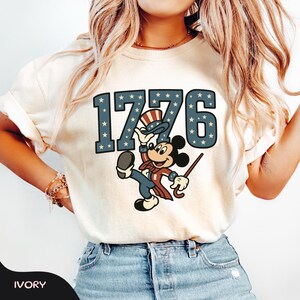 May include: Ivory-colored t-shirt featuring the number "1776" in blue with white stars. A cartoon character in patriotic attire is holding a cane and a hat. The word "IVORY" is at the bottom of the shirt.