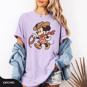 May include: A light purple t-shirt featuring a cartoon character dressed as a cowgirl, holding a lasso. The character wears a brown hat, a red scarf, and a pink dress with white polka dots. The word "ORCHID" is at the bottom.