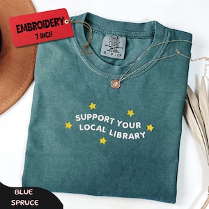 May include: Blue spruce t-shirt with the text "Support Your Local Library" and yellow star accents. The shirt has a round neckline and short sleeves. A gold necklace with a pendant is draped on the shirt. A red tag reads "Embroidery 7 inch".
