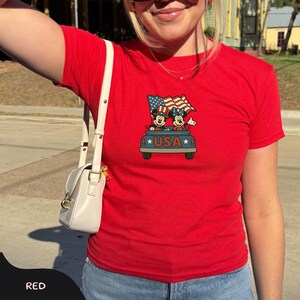 May include: A red t-shirt featuring a graphic of Mickey and Minnie Mouse in a blue truck with a waving American flag. The word "USA" is printed on the truck. The shirt is short-sleeved.