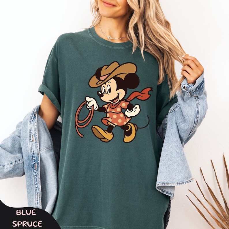 May include: Teal-colored t-shirt featuring a cartoon Minnie Mouse dressed as a cowgirl, holding a lasso. Minnie wears a brown cowboy hat, a red scarf, and a pink dress with orange polka dots. The text "BLUE SPRUCE" is at the bottom.