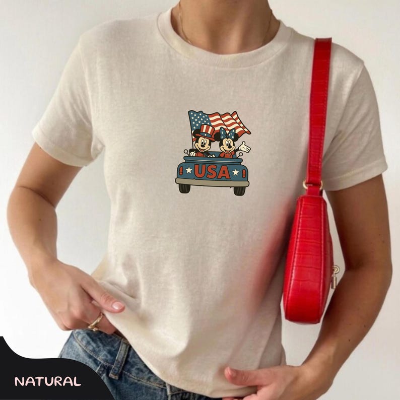 May include: Cream-colored t-shirt featuring a graphic of Mickey and Minnie Mouse in a blue truck with American flags and the word "USA". The shirt is paired with blue jeans and a red purse.