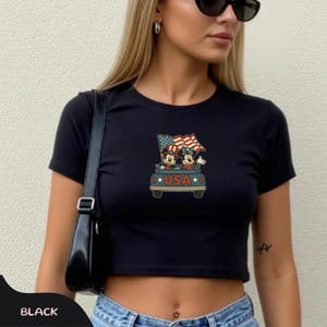 May include: Black crop top featuring a graphic of Mickey and Minnie Mouse in a car with American flags and the word "USA". The shirt has short sleeves and a crew neck.