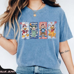 May include: Blue jean colored t-shirt featuring four cartoon characters in patriotic designs. Donald Duck is wearing a red and white hat. Mickey Mouse holds a torch. Goofy is sitting on a barrel. Pluto is holding a flag.