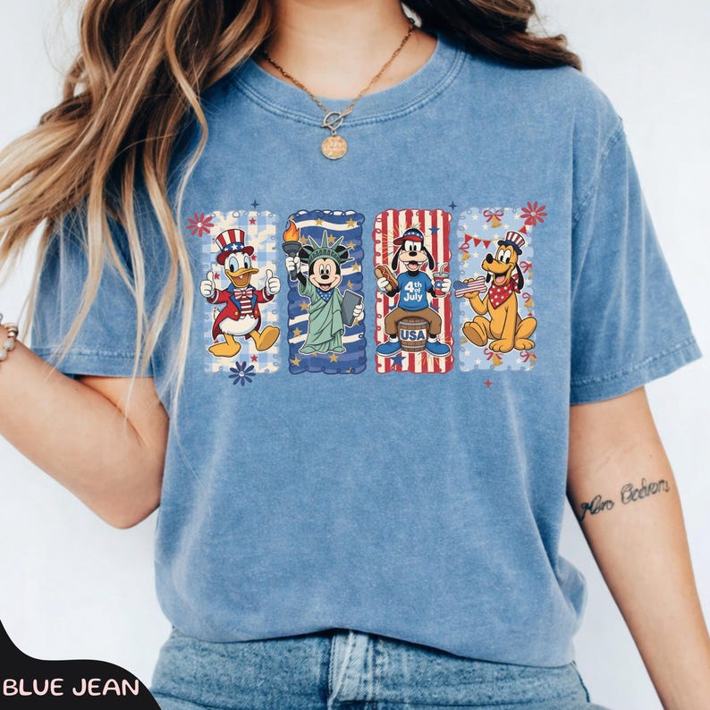 May include: Blue jean colored t-shirt featuring four cartoon characters in patriotic designs. Donald Duck is wearing a red and white hat. Mickey Mouse holds a torch. Goofy is sitting on a barrel. Pluto is holding a flag.