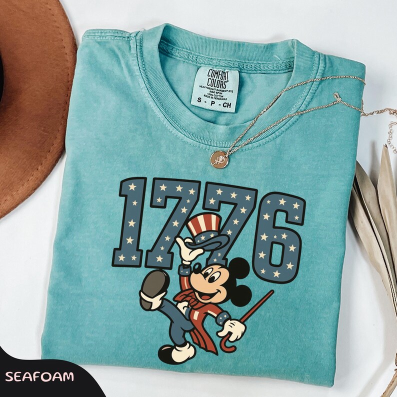 May include: Seafoam green t-shirt with the text "1776" in navy blue with white stars. The shirt features a cartoon character in patriotic attire. A gold necklace and a brown hat are visible.