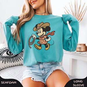 May include: Seafoam green long-sleeve sweatshirt featuring a cartoon Minnie Mouse dressed as a cowgirl, holding a lasso. Minnie Mouse is wearing a brown cowboy hat, a red scarf, and a polka-dot dress. The sweatshirt has the word "Seafoam" printed on the bottom.
