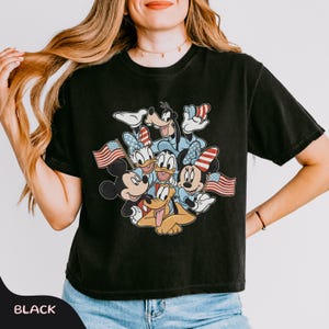 May include: Black t-shirt featuring a graphic of Mickey Mouse, Minnie Mouse, Donald Duck, Daisy Duck, Goofy, and Pluto, each holding an American flag. The characters are in a patriotic design. The word "BLACK" is at the bottom.