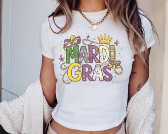 May include: White t-shirt with a colorful "Mardi Gras" graphic. The design features a crown, mask, and beads in green, purple, and yellow. The shirt is short-sleeved and has a crew neck.