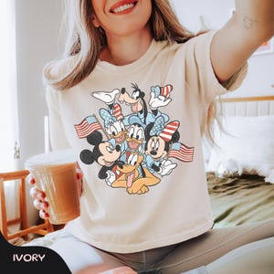 May include: Ivory-colored t-shirt featuring a graphic of Mickey Mouse, Minnie Mouse, Donald Duck, Daisy Duck, Goofy, and Pluto, each holding an American flag. The characters are in patriotic attire. The word "IVORY" is printed at the bottom.