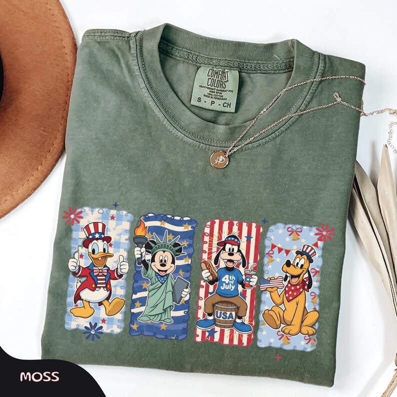 May include: Moss green Comfort Colors t-shirt featuring four cartoon character designs with patriotic themes. Donald Duck, Mickey Mouse, Goofy, and Pluto are depicted in red, white, and blue attire, celebrating the 4th of July. A gold necklace is draped across the shirt.