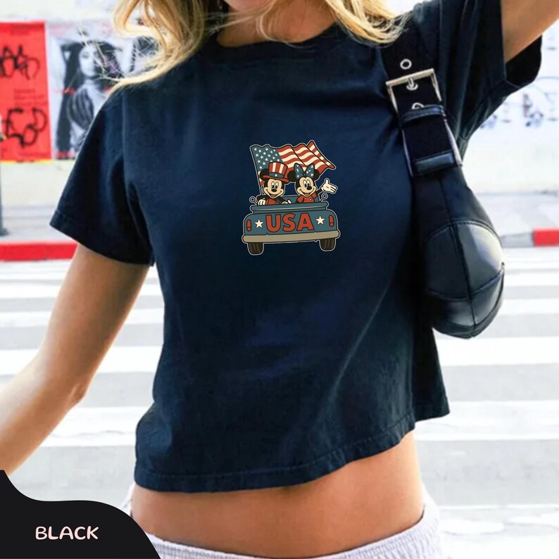 May include: Black crop top featuring a graphic of Mickey and Minnie Mouse in a truck with American flags and the word "USA". The design is in red, white, and blue. The shirt has short sleeves.